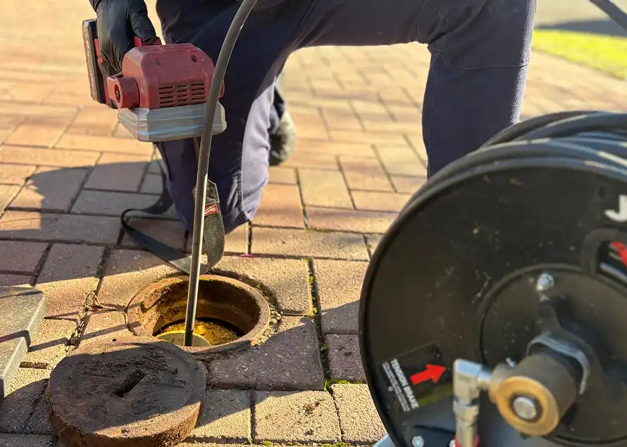 A plumber feeding a powered drain machine cable down through an open inspection point in a paved driveway, with the jetter hose reel alongside.