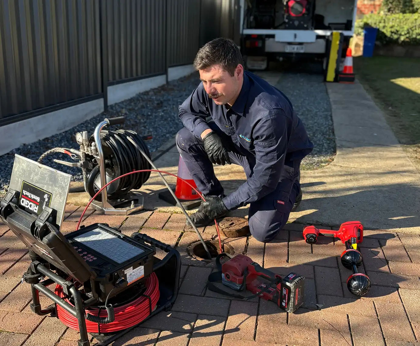 Marc from Allmat Plumbing kneeling beside an open inspection point, feeding a RIDGID SeeSnake drain camera into the drain with the reel and locator on the paving.