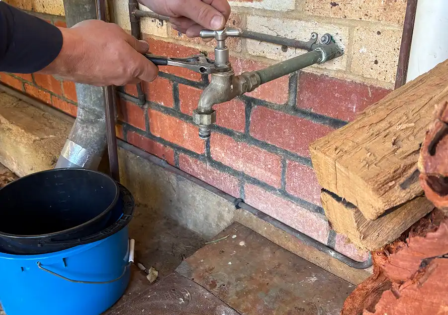 Hands using a shifter on the brass body of an outdoor garden tap mounted to a brick wall, with a bucket underneath to catch the water.