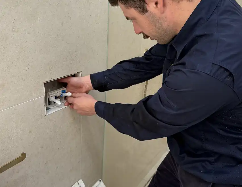 A plumber reaching into the open flush plate recess of a concealed in-wall toilet cistern in a tiled bathroom.