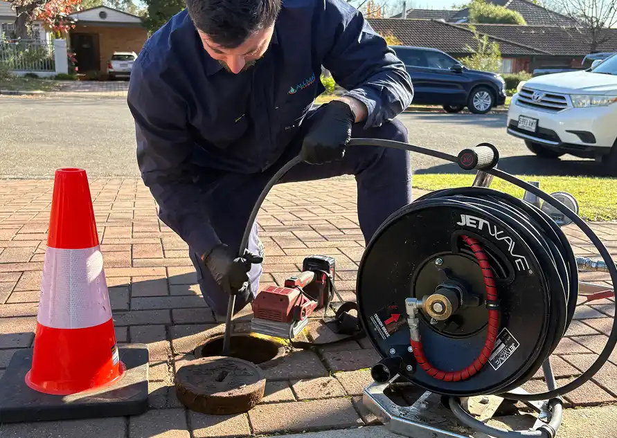 A plumber kneeling at an open inspection point on a paved driveway, feeding a jetter hose into the drain from a Jetwave hose reel.